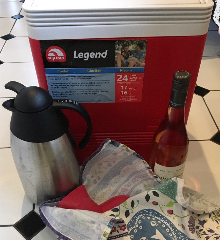 Red Igloo cooler with coffee carafe, bottle of wine, and red rubber hot water bottle wrapped in towel