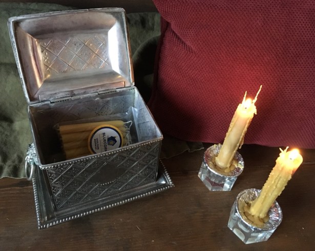 2 lit tapers on wooden table next to open silver chest containing beeswax candles