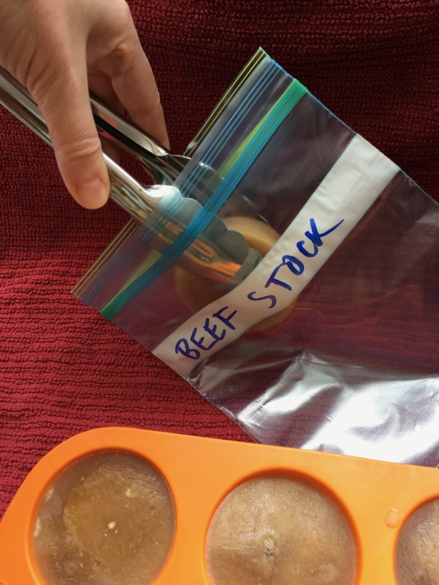 Hand holding stainless steel tongs placing frozen puck of broth into baggie labeled Beef Stock