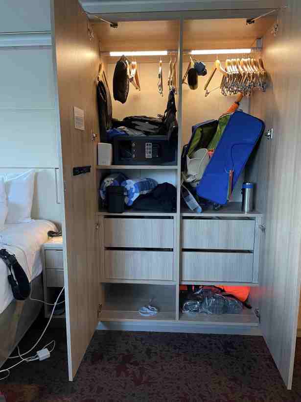 Closet next to bed with haphazard arrangement of items, some still in suitcase