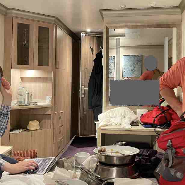 4 people shown in disorderly inside room; 3 sit on beds while 4th stands near tv wall