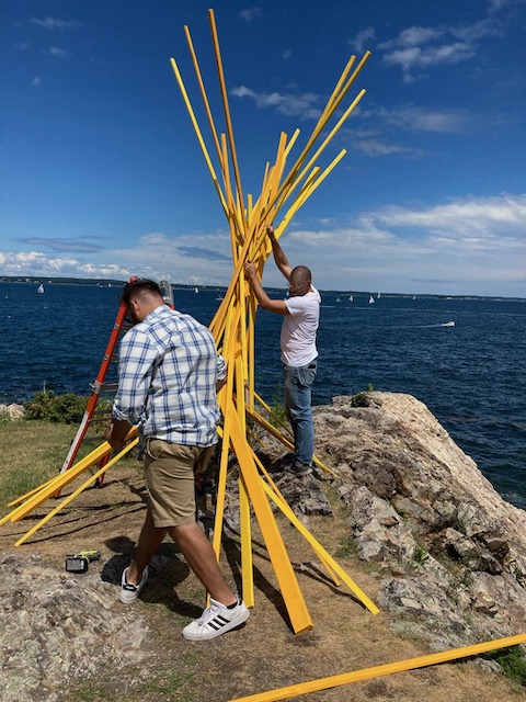 Two men assemble yellow sticks into teepee formation with sailboats visible on ocean behind them