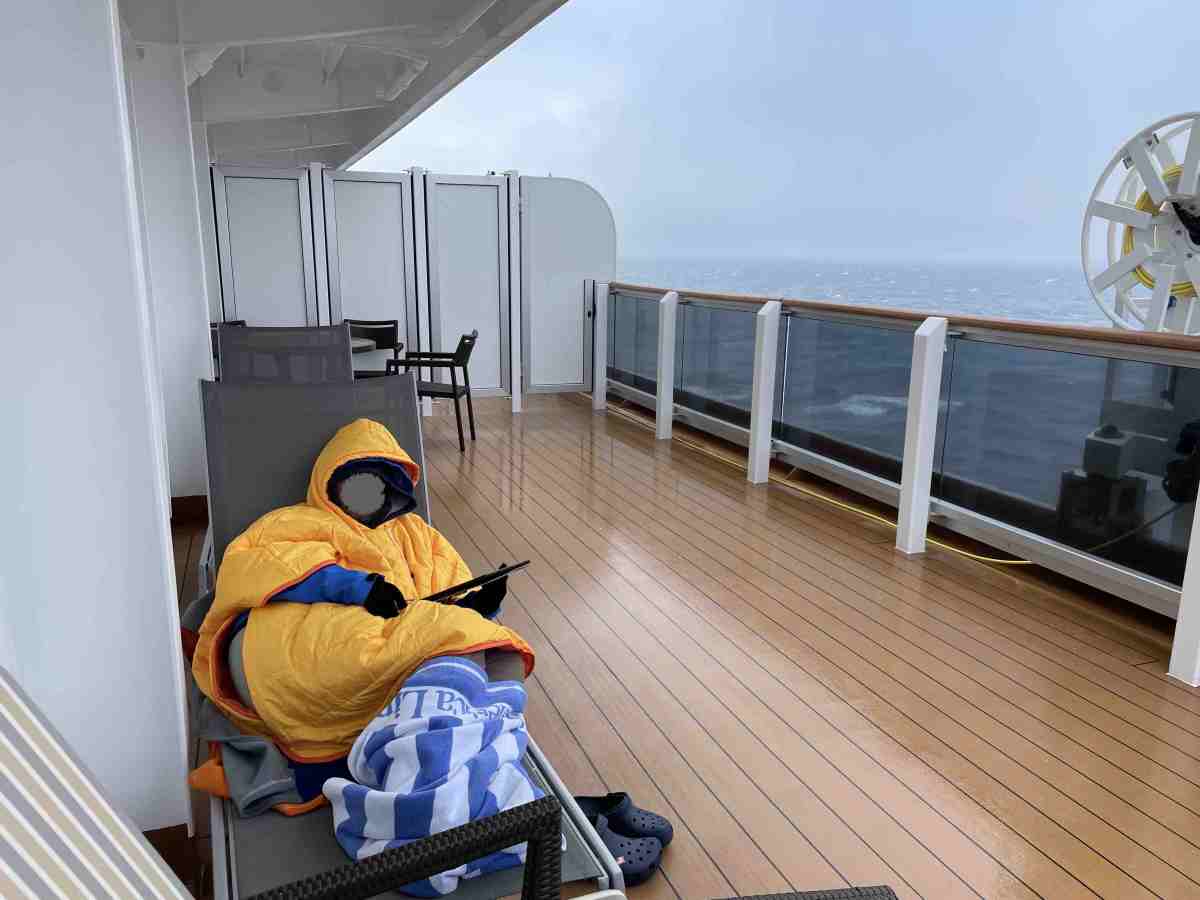 Man on wet cruise ship balcony with puffy poncho on body and beach towel wrapped around feet while working on tablet
